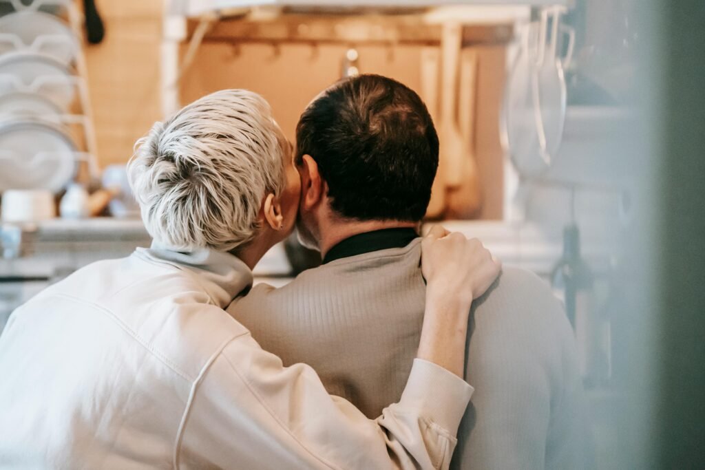 Back view of unrecognizable couple sitting closely while cuddling and caressing cheeks gently in kitchen